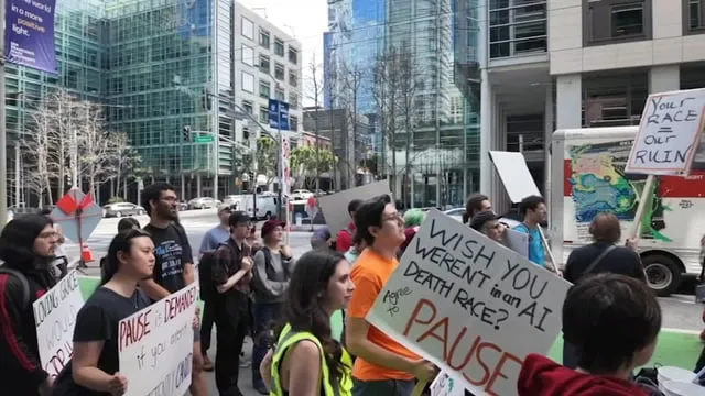 Demonstranter i San Francisco håller skyltar med texter som "PAUSE" och "AI DEATH RACE". Höga byggnader i bakgrunden.