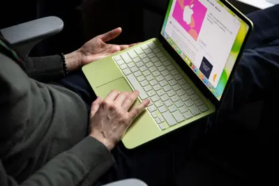 En person sitter med en limegrön MacBook i knät och skriver. Skärmen visar en artikel om Apple.