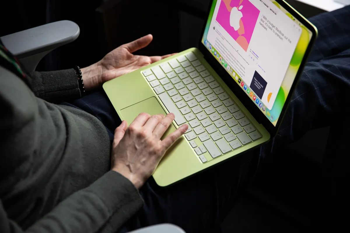 En person sitter med en limegrön MacBook i knät och skriver. Skärmen visar en artikel om Apple.