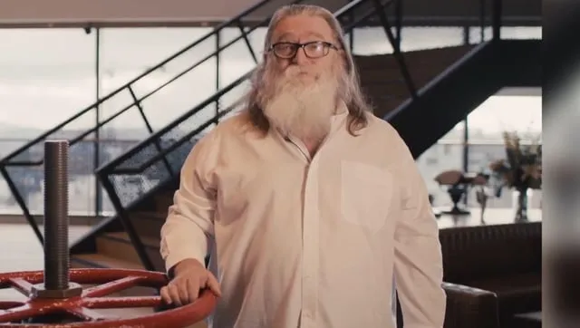 Gabe Newell med långt vitt skägg och glasögon står framför en röd ventil. Trappa i bakgrunden.