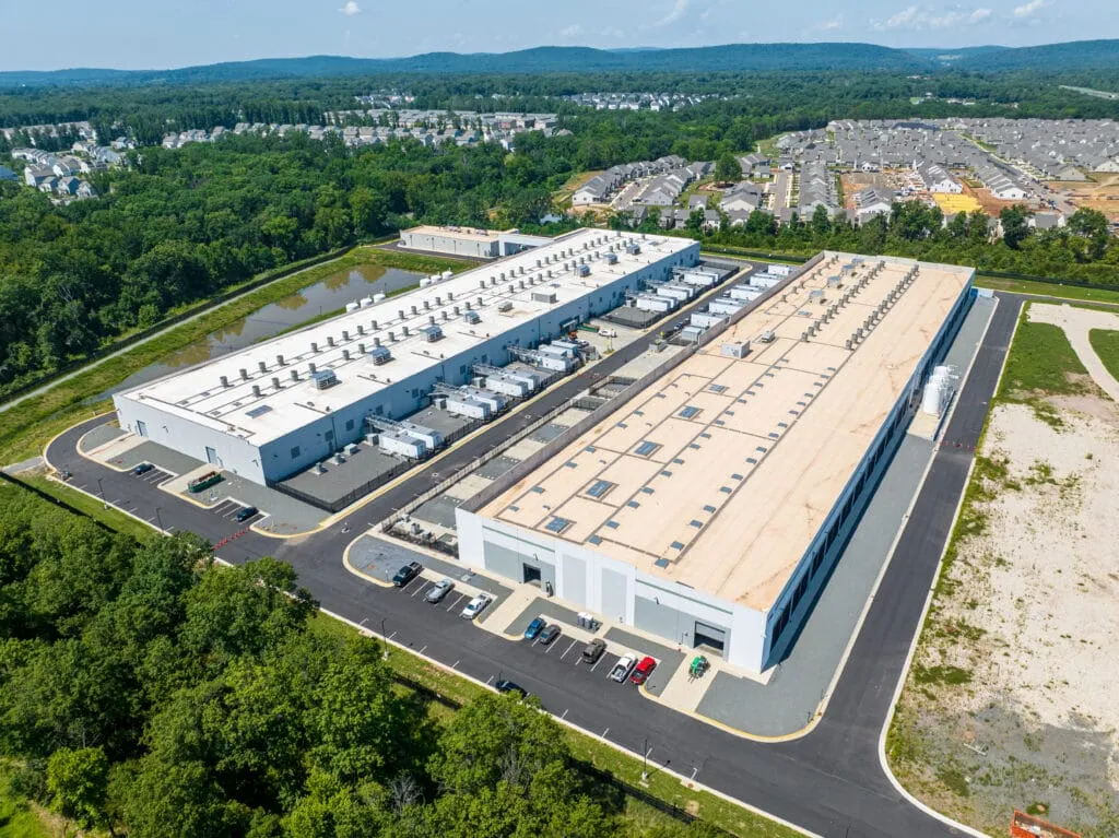 Flygfoto av två stora, grå byggnader med fläktar på taken, omgivna av grönska och bostadsområden.