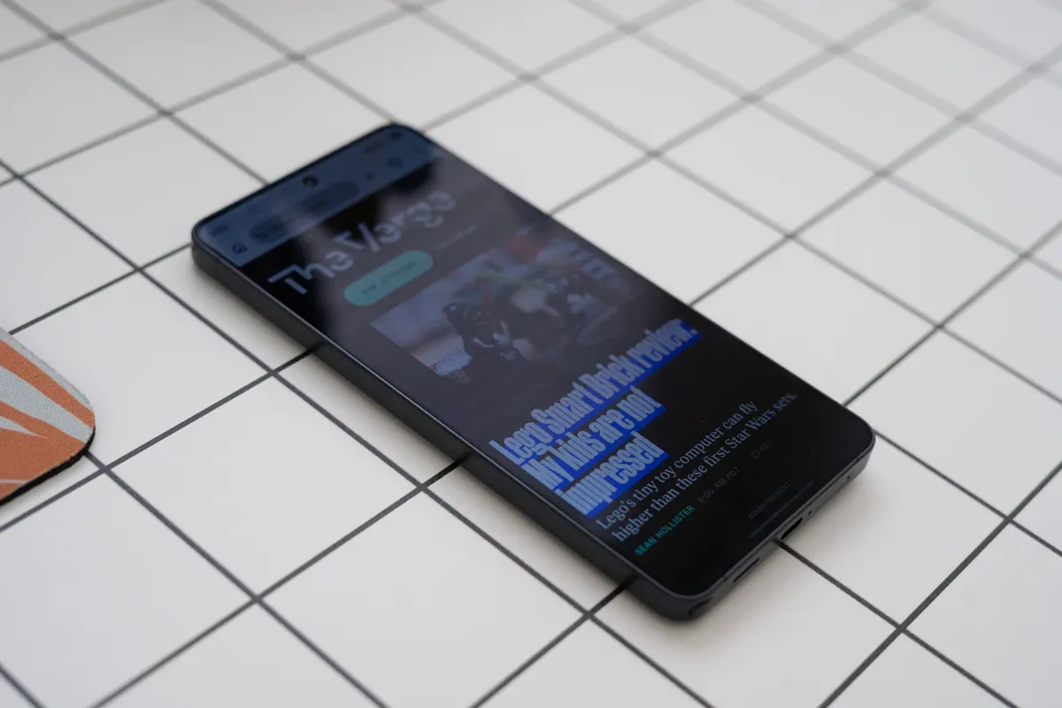 En svart mobiltelefon visar en artikel på The Verge med rubriken "Lego Smart Brick review". Mobilen ligger på ett rutigt underlag.