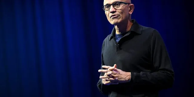 Satya Nadella, Microsofts VD, står framför en blå bakgrund. Han bär glasögon och en mörk tröja.