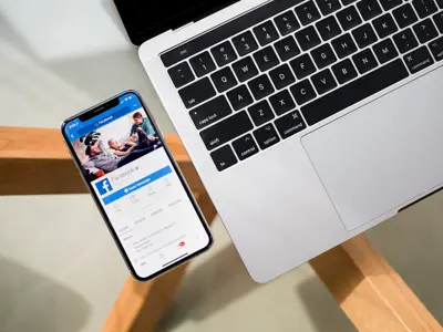 En iPhone visar Facebook-appen bredvid en öppen laptop på ett bord.