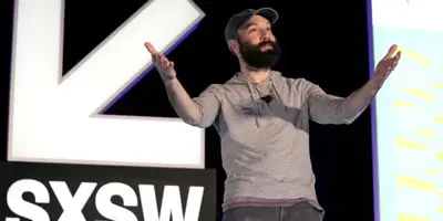 En man med skägg och keps står på en scen med SXSW-loggan i bakgrunden. Han gestikulerar med händerna.