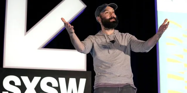 En man med skägg och keps står på en scen med SXSW-loggan i bakgrunden. Han gestikulerar med händerna.
