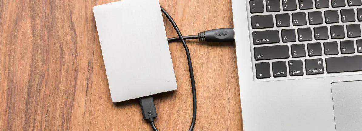 En extern hårddisk är ansluten till en bärbar dator med en USB-kabel. Båda ligger på ett träbord.