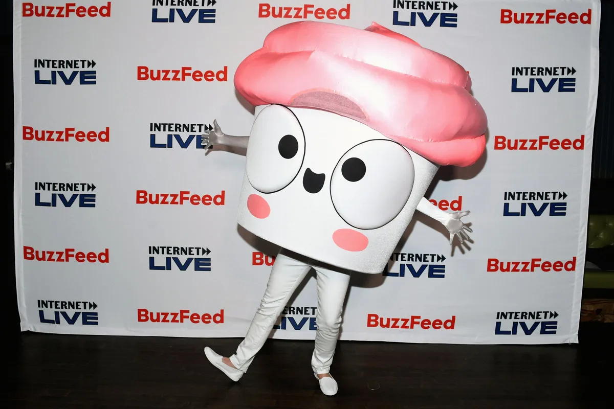 En person i en cupcake-maskot står framför en Buzzfeed-bakgrund. Maskoten är vit med rosa frosting.