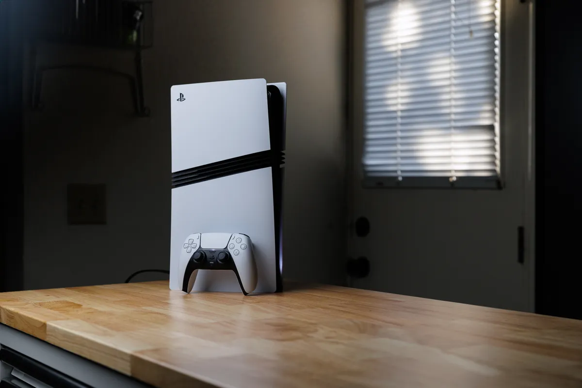 En Playstation 5-konsol och en handkontroll står på en träbänk. I bakgrunden syns en dörr med persienner.