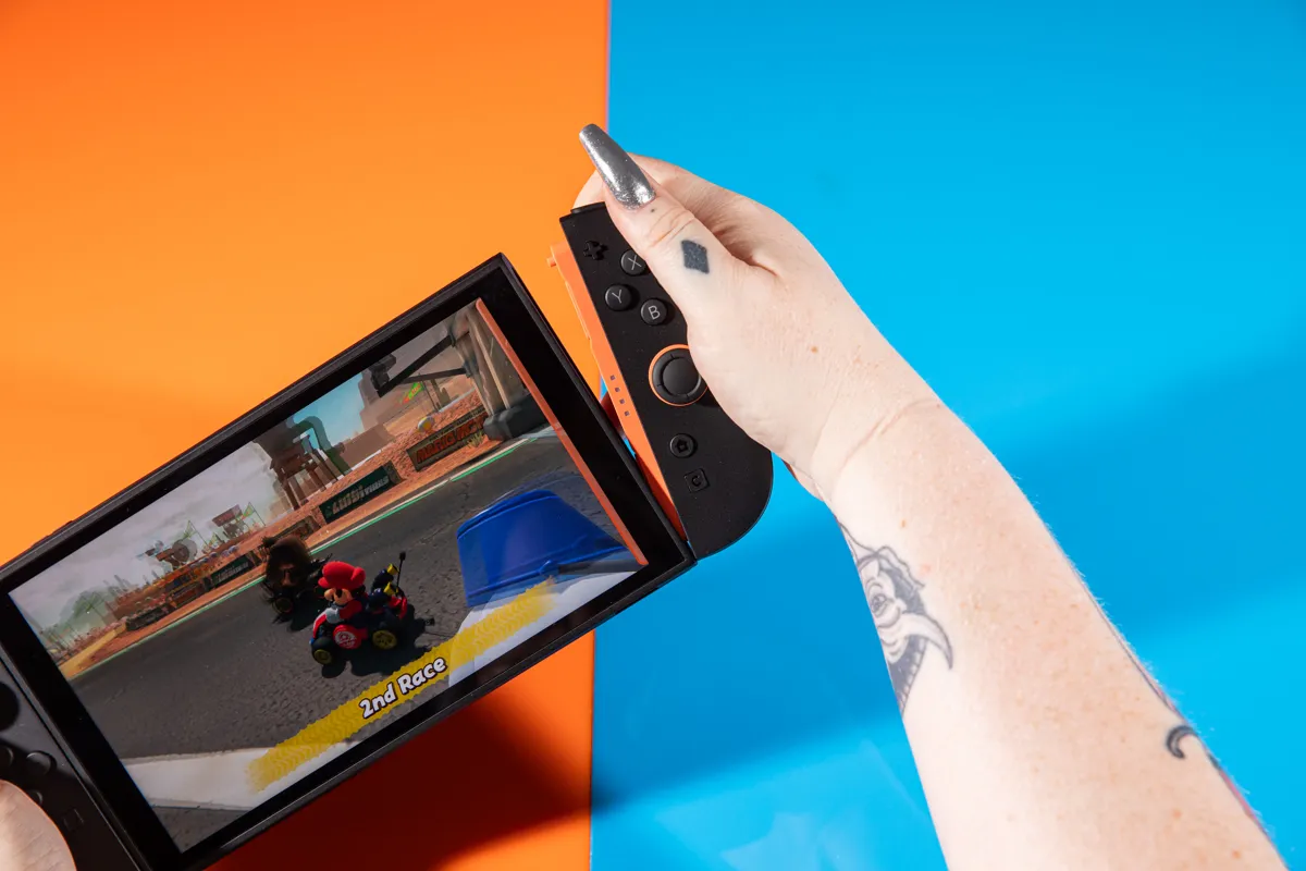 En person spelar Mario Kart på en Nintendo Switch med orange Joy-Cons. Bakgrunden är orange och blå.