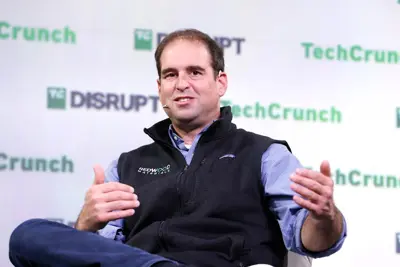 En man i väst och skjorta sitter framför en TechCrunch-bakgrund.