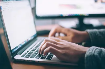 En person skriver på en laptop. En datorskärm syns i bakgrunden.